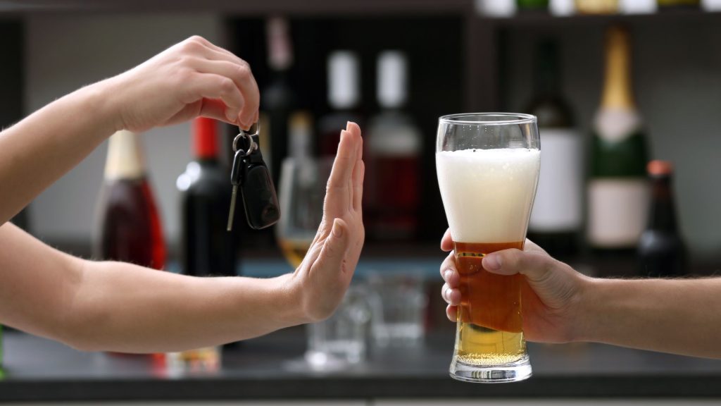 A hand is shown refusing a glass of alcohol while holding a set of car keys. 